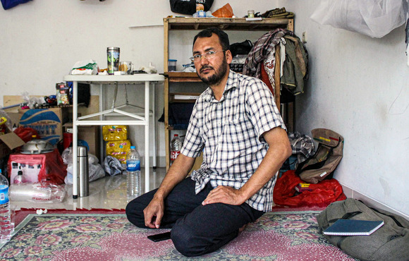 A man kneels on a cement floor in front of a makeshift shelter. 