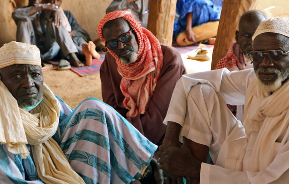 Three men sit on the ground under a shelter, looking up at the camera. 