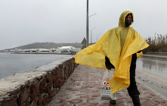 A woman walks along a coastal promenade wearing a yellow rain poncho, and holding a plastic grocery bag. Her face is covered by a mask. 