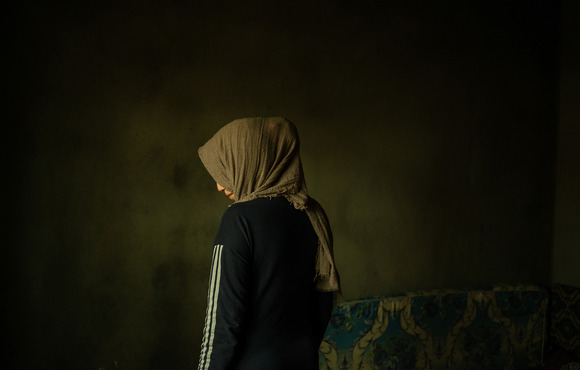 An image of a woman in a headscarf standing in a near empty room from behind, looking slightly down and to the side.