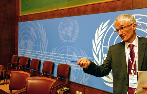 UN humanitarian chief Mark Lowcock stands to the right of the image, looking past the camera and holding out a pen in his hand. Empty seats are pictured in the background, as well as a large poster of the UN's brand.