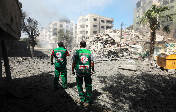 Two medics dressed in green uniforms stand in front of demolished buildings with their backs to the camera.