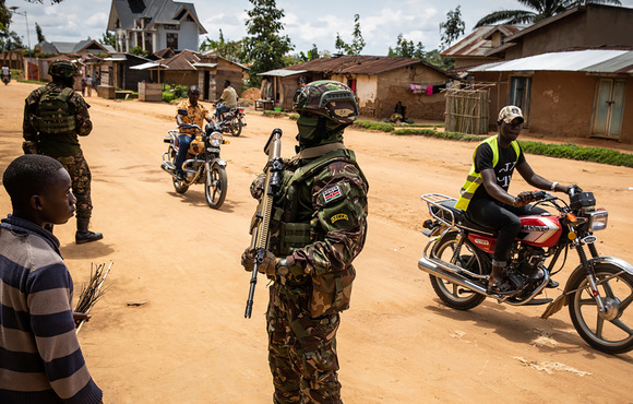 Several armed soldiers stand on a large road lined by small dwellings.