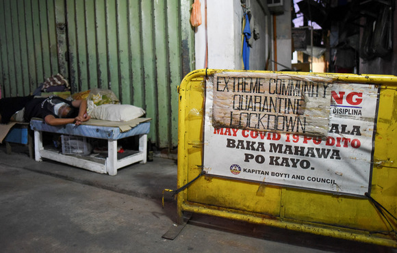 A sign blocks a city street indicating a local lockdown – in the background of the image, a figure lies sideways on a bench.