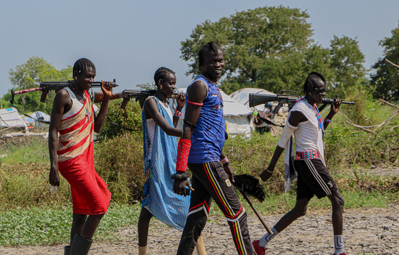 4 armed youth walk along a path lined by grass, one looks at the camera with a smile. 