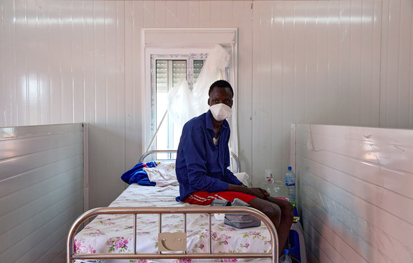 The image shows a young man sitting on a wrought iron hospital bed – a window half-covered by a curtain is above the bed