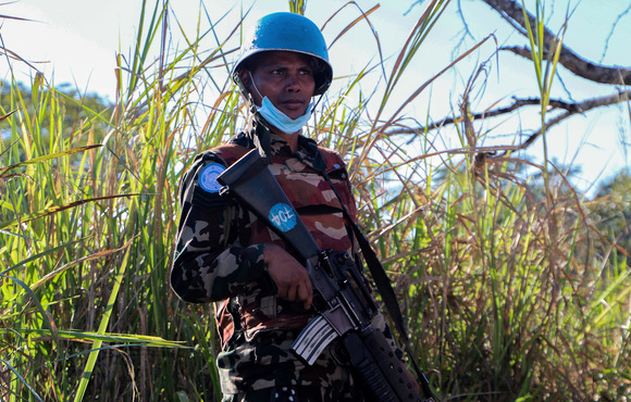A woman in a blue UN helmet holds a rifle in front of a green field. 