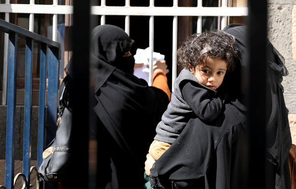 A boy being held by his mother, who is waiting for foodstuff assistance at a window counter, looks at the camera through an iron bar fence. 