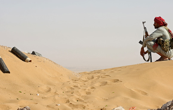 A man crouches on a sand dune, holding a rifle
