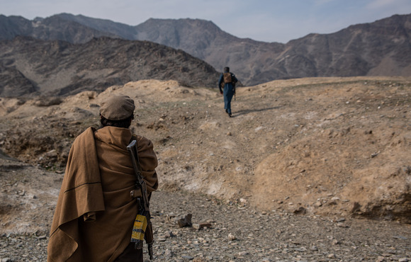 Two figures with weapons trek across arid mountains.